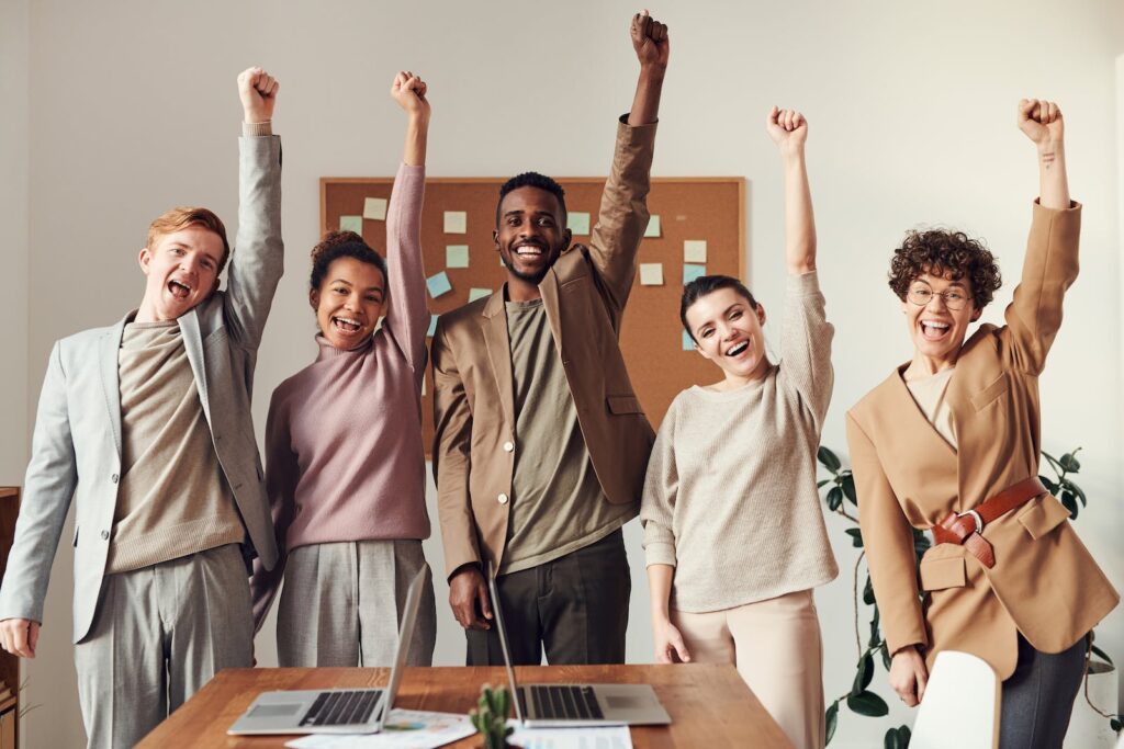 Un groupe de gens d'affaires levant les bras au bureau, démontrant leur enthousiasme et leur esprit d'équipe dans un environnement de travail animé.