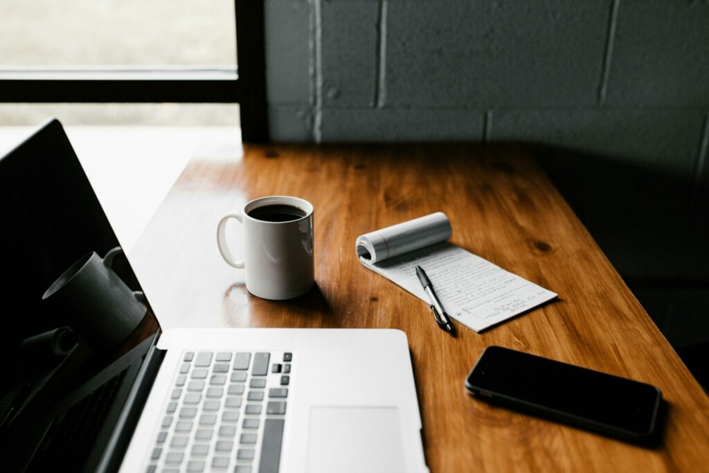Un bureau en bois avec un ordinateur portable ouvert exécutant un logiciel de recrutement, une tasse de café, un bloc-notes avec des notes manuscrites pour planifier l'entretien, un stylo et un smartphone près d'une fenêtre.