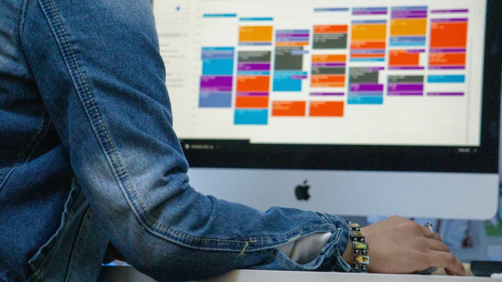 Une personne portant une veste en jean est assise à un bureau, utilisant un iMac pour planifier l'entretien avec un calendrier numérique à code couleur, mettant en valeur la gestion efficace des entretiens ATS.