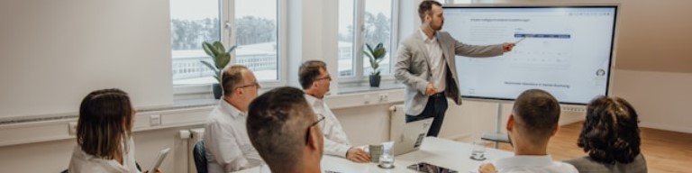 Dans une salle de conférence moderne, un consultant en blazer présente des données RH et juridiques sur un grand écran à un groupe de collègues assis. Photos : ATS dans le secteur du conseil en management, juridique, comptable et RH pour améliorer l'expérience candidat