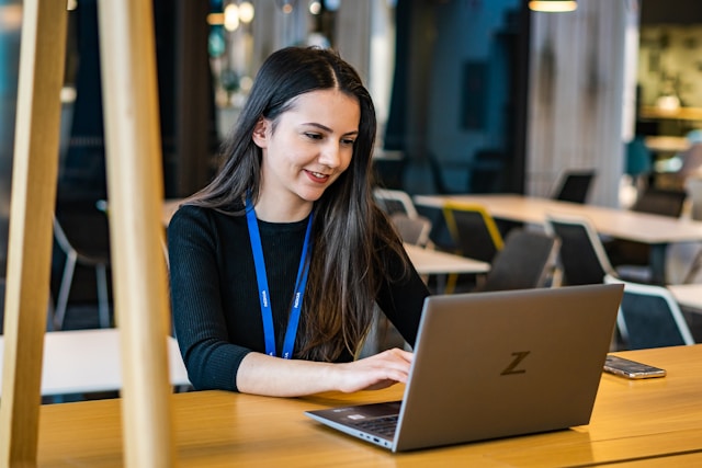 Une recruteuse aux longs cheveux bruns, portant un cordon bleu, est assise à une table dans un bureau moderne, travaillant sur un ordinateur portable, naviguant efficacement dans un logiciel de recrutement ATS. Image d'illustration : Améliorer l'expérience candidat dans Le BTP et la construction