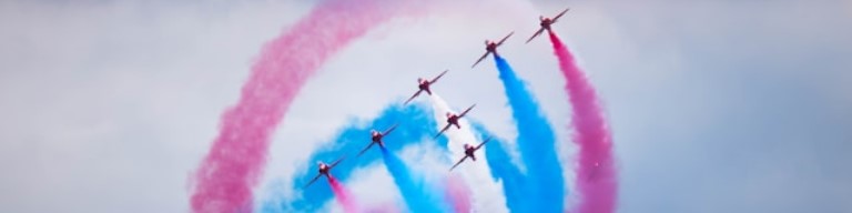 Six avions volent en formation, laissant des traînées de fumée rose et bleue dans un ciel nuageux, illustrant la précision et le travail d'équipe valorisés dans la défense et essentiels pour un recrutement réussi. Image d'illustration pour expliquer comment un ATS de recrutement aide le secteur de l'aéronautique et de la défense pour améliorer l'expérience candidat