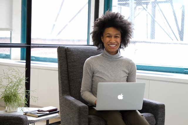 Une personne, peut-être un chargé de recrutement, est assise dans un fauteuil gris, souriant tout en utilisant un ordinateur portable. Une petite plante et des cahiers sont posés sur une table à côté d'eux. De grandes fenêtres se trouvent à l'arrière-plan. Illustration d'un guide pour Informatique concernant : Trouver des candidats gratuitement