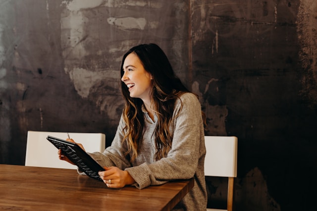 Une femme aux cheveux longs est assise à une table en bois, souriante et tenant une tablette, vêtue d'un pull gris. Elle est peut-être à la recherche d'opportunités d'emploi ou de conseils en matière de recrutement de la part d'experts de la DRH. Contexte BTP - Organiser des entretiens structurés