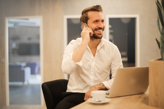 Un recruteur en chemise blanche est assis à un bureau avec un ordinateur portable, souriant tout en parlant au téléphone. Une tasse à café et une plante sont posées sur le bureau. Image illustrant : Informatique - Améliorer l’expérience candidat