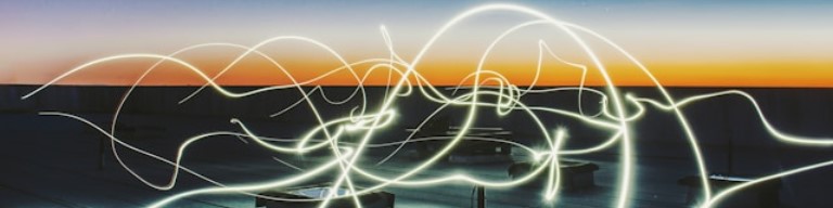Des traînées courbes de lumière blanche apparaissent au-dessus d'un toit au crépuscule, avec un ciel orange et bleu clair en arrière-plan, donnant à la scène une impression d'énergie vibrante. Image abstraite pour promouvoir l'usage d'un ATS dans le secteur de l'énergie pour centraliser les offres et candidatures