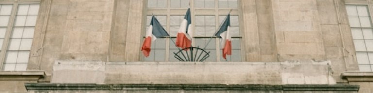 Trois drapeaux français sont déployés au-dessus de l'entrée d'un bâtiment en pierre doté de grandes fenêtres, ce qui laisse supposer qu'il s'agit d'une importante institution du secteur public, peut-être impliquée dans le recrutement ou les tâches administratives, utilisant un ATS. Image d'illustration : logiciel ATS contribuant à centraliser les offres et candidatures dans le secteur public et la fonction publique.