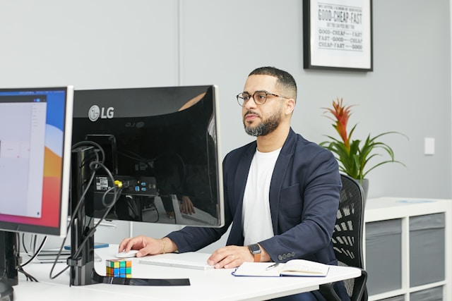 Un homme à lunettes, peut-être un chargé de recrutement, est assis à un bureau et travaille sur un ordinateur, avec un carnet, un stylo et un Rubik's cube à proximité. Une citation motivante encadrée et une plante ajoutent de l'inspiration à son espace de travail. Pour illustrer : Informatique avec Rédiger une annonce de recrutement attractive