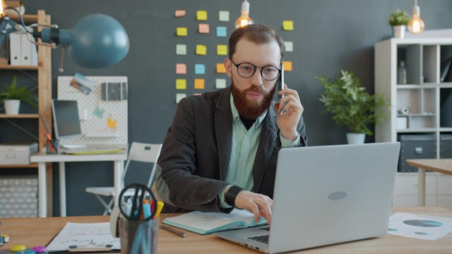 Un homme à lunettes travaille sur un ordinateur portable à un bureau, tout en parlant au téléphone - peut-être pour discuter d'un ATS ou d'un logiciel de recrutement - avec des notes autocollantes colorées couvrant le mur en arrière-plan. Photo d'illustration - Améliorer l'expérience candidat dans La santé