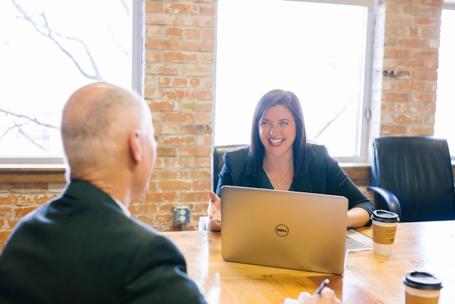 Une DRH est assise à une table avec un ordinateur portable, souriant et discutant d'un logiciel de recrutement avec un recruteur en face d'elle dans un bureau lumineux aux murs de briques et aux grandes fenêtres. Illustration : Améliorer l'expérience candidat pour Le monde associatif