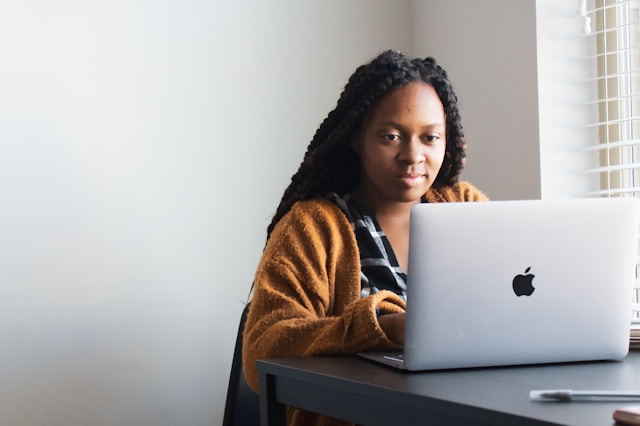 Une Recruteuse est assise à un bureau et utilise un ordinateur portable Apple argenté près d'une fenêtre avec des stores, examinant probablement des candidats par le biais de son logiciel de recrutement. Elle porte un cardigan marron et une chemise noire et blanche. Image illustrant : Centraliser les offres et candidatures pour Le monde associatif