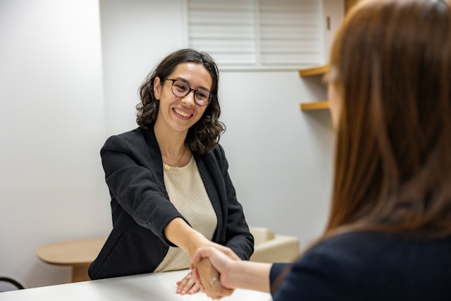 Deux femmes sont assises autour d'une table dans un bureau, souriantes et se serrant la main, suggérant une réunion de recrutement réussie entre un drh et un chargé de recrutement. Illustration de énergie - Rédiger une annonce de recrutement attractive
