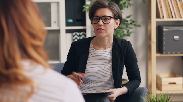 Une personne aux cheveux courts et aux lunettes, tenant un stylo et un bloc-notes, discute avec une autre personne des possibilités d'emploi dans un bureau - peut-être pour préparer un entretien avec un drh ou un chargé de recrutement. Article pour Informatique sur Améliorer sa marque employeur pour attirer les talents