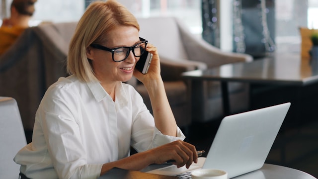 Une femme portant des lunettes est assise à une table, souriante, tout en parlant à un recruteur sur son smartphone et en utilisant un ordinateur portable dans un café moderne et lumineux. Environnement contextuel : énergie - Trouver des candidats gratuitement