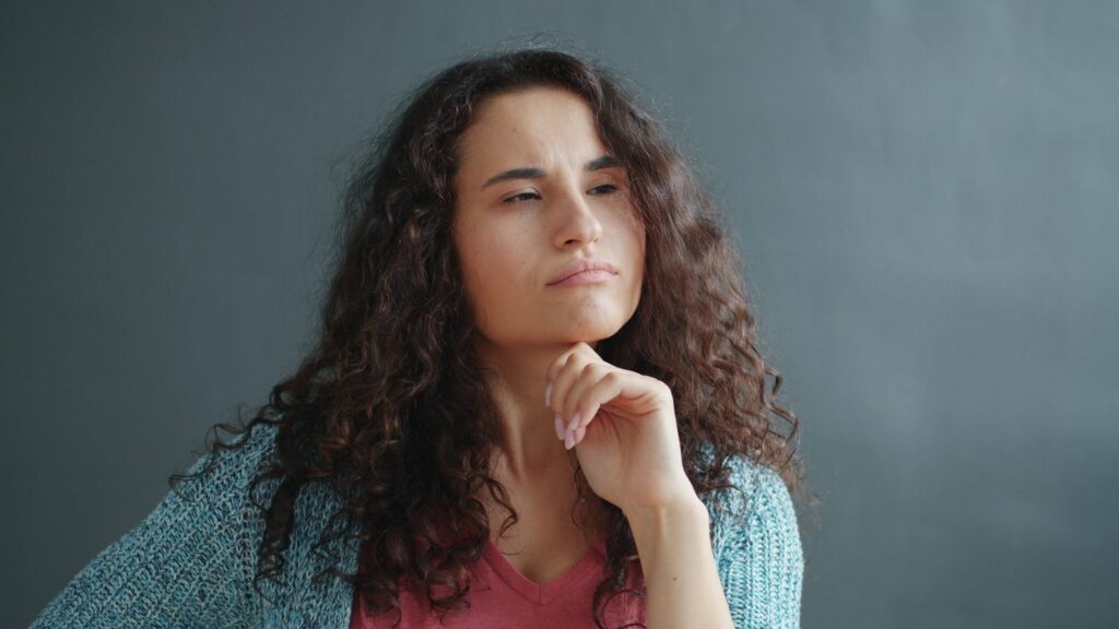 Femme aux cheveux bouclés, le menton posé sur la main, le regard pensif tourné vers le côté, sur un fond uni, comme si elle réfléchissait à l'utilité que l'ATS apporte à son quotidien.